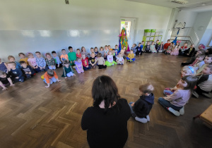 Dzieci siedzą w kole na sali gimnastycznej, słuchają instrukcji przekazywanych przez nauczyciela