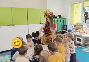 Pani Jesień trzyma w ręku parasolkę, w której są liście i pokazuje ją dzieciom