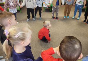 Dzieci stoją w kole, jeden chłopiec kuca w środku koła