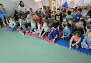 Dzieci siedzą na sali gimnastycznej, niektóre zgłaszają się do odpowiedzi