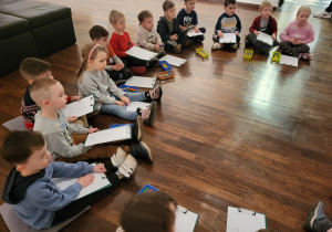 Dzieci siedzą w kole i trzymają podkładki z kartkami