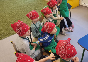 Dzieci w chustkach i fartuszkach siedzą obok stolików