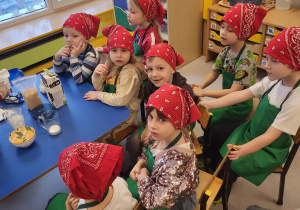 Dzieci w chustkach i fartuszkach siedzą obok stolików