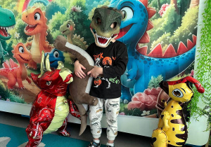 Chłopiec w masce prezentuje swojego pluszowego dinozaura trzymając go w obu dłoniach