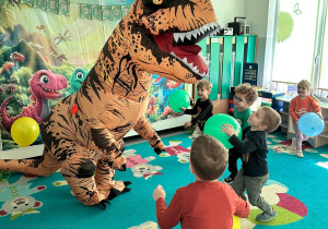 Dzieci uczestniczą we wspólnej zabawie ruchowej z dmuchaną postacią dinozaura